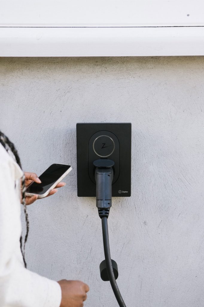 Elbillader montert på vegg med tilkoblet kabel. Kvinne står med telefon i hånden.
